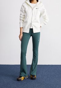 Vit zip-up hoodie med dragsko, tillsammans med mörkgröna utsvängda träningsbyxor. Svart Nike-logotyp på både tröjan och byxorna, samt texturerade skor.