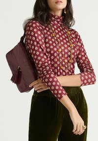 Blusa color bordeaux con motivo geometrico, abbinata a pantaloni di velluto verde. Viene tenuta una borsa di colore marrone scuro con dettaglio a nappina.