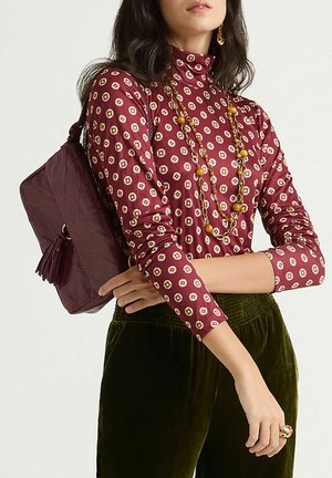 Blusa color bordeaux con motivo geometrico, abbinata a pantaloni di velluto verde. Viene tenuta una borsa di colore marrone scuro con dettaglio a nappina.