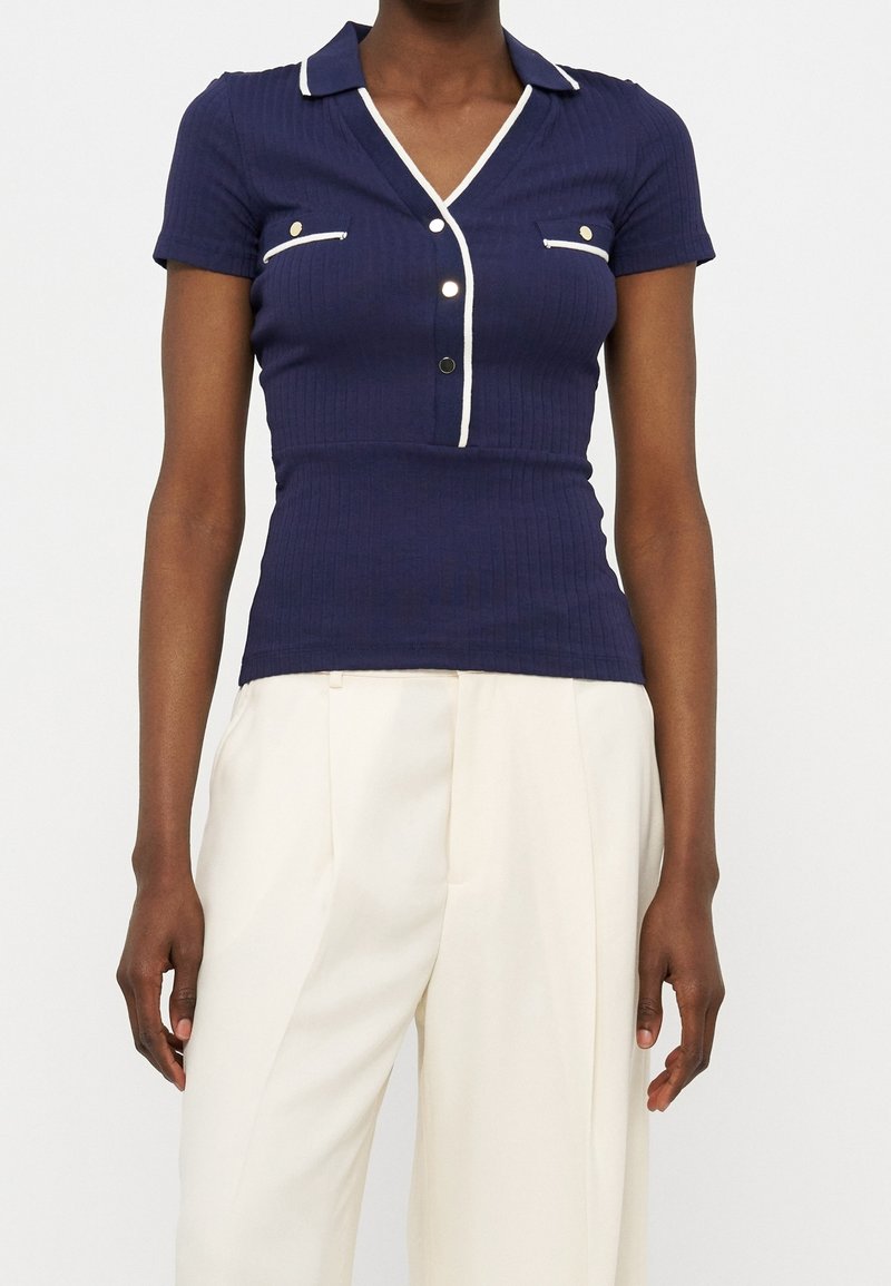 Femme portant un polo côtelé bleu marine à manches courtes avec un liseré blanc et un pantalon blanc plissé taille haute sur fond neutre.