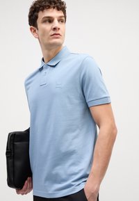 Gai zilas polo krekls ar klasisku apkakli, īsām piedurknēm, teksturētu audumu un mazu iegrieztu logo uz krūtīm. Tur rokās melnu somu.