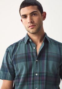 Kortærmet ternet skjorte i blågrøn, grøn og lilla med et struktureret mønster, der har en skjortekrave med knapper og hvide knapper.