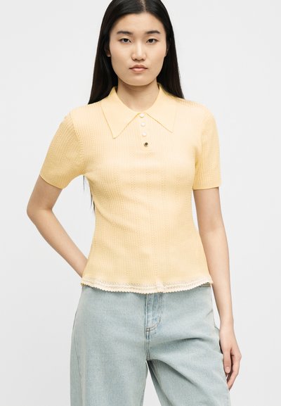Νεαρή γυναίκα με κοντομάνικο πλεκτό πόλο μπλουζάκι σε απαλό κίτρινο χρώμα με κουμπιά και ανοιχτό μπλε τζιν, στέκεται με το ένα χέρι στη μέση.