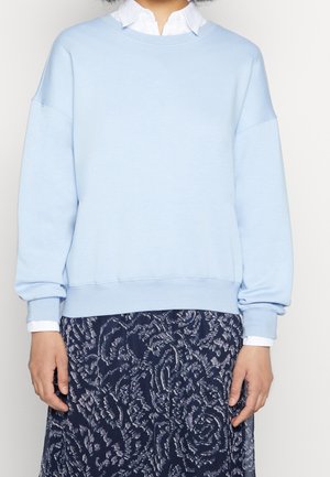 Personne portant un sweat-shirt bleu clair sur une chemise blanche à col et une jupe bleu marine à motif floral blanc, debout devant un fond uni.