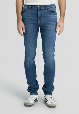 Mężczyzna ubrany w niebieskie, dopasowane jeansy i białe trampki z czarnymi paskami, stojący na jednolitym jasnym tle.