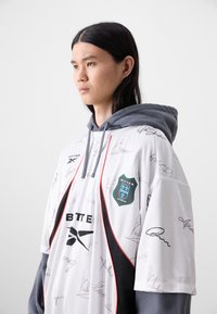 White oversized jersey with gray hood, featuring black accents, colorful logo, and multiple signatures printed across the fabric.