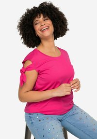 Rosa T-Shirt mit kurzen Ärmeln und verdrehtem Knoten-Detail an den Schultern, kombiniert mit hellblauen Jeans mit mehrfarbigen Schmucksteinelementen.