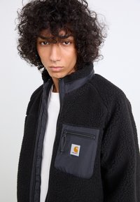 Sort fleecejakke med en lynlåslomme. Har en kontrasterende nylon frontpanel og et Carhartt-logo på lommen. Krøllet hår synligt.