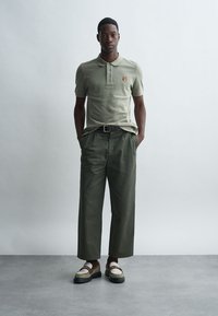 Ανοιχτό ελ olive πράσινο polo μπλουζάκι με κεντημένο μπάλωμα, συνδυασμένο με φαρδιά ελ olive παντελόνια, που τονίζονται από μια μαύρη ζώνη και κομψά loafers.