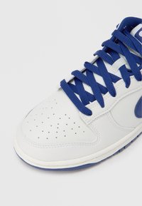 Biały skórzany sneaker z granatowymi sznurowadłami i akcentami. Posiada perforacje na palcach dla wentylacji. Płaska podeszwa.