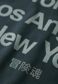 Tela gris oscuro con texto grande en gris claro que dice "Los Ángeles" y "Nueva York", con caracteres adicionales en una fuente más pequeña debajo.