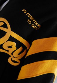 Svart sweatshirt med tydlig gul text och en stor skrivstil. Har två horisontella gula ränder på ärmarna.