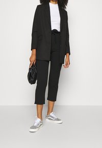 Svart oversized blazer över en vit t-shirt, kombinerad med svarta cropped-byxor och grå sneakers. Svart handväska med samlad design.