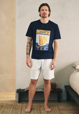 Muž nosiaci tmavomodré tričko s motívom ryby a hranoliek a biele krátke nohavice, stojaci bosý na koberci v minimalistickej spálni.