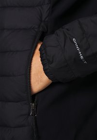 Svart puffjacka med en ficka med dragkedja, elastiska ärmslut och "OMNI-HEAT"-märke. Materialet har en slät textur och vadderad design.