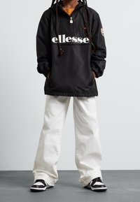 Zwarte windjack met het "ellesse" logo over de voorkant, half-ritssluiting bij de kraag, elastische manchetten, gecombineerd met witte wijde broek en zwarte sneakers.