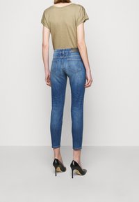 Woman wearing fitted blue jeans, beige short-sleeve top, and black high-heeled shoes standing against a plain light background.