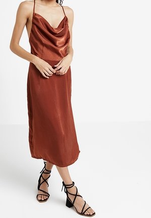 Femme portant une robe midi en satin couleur rouille avec fines bretelles et col bénitier, associée à des sandales noires à talons blocs et lanières.