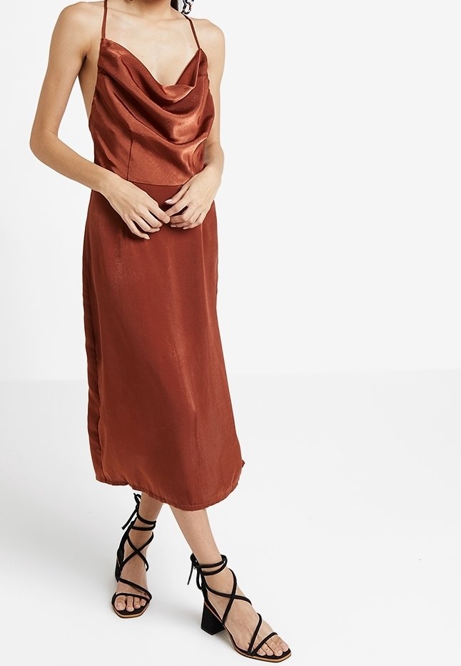 Femme portant une robe midi en satin couleur rouille avec fines bretelles et col bénitier, associée à des sandales noires à talons blocs et lanières.