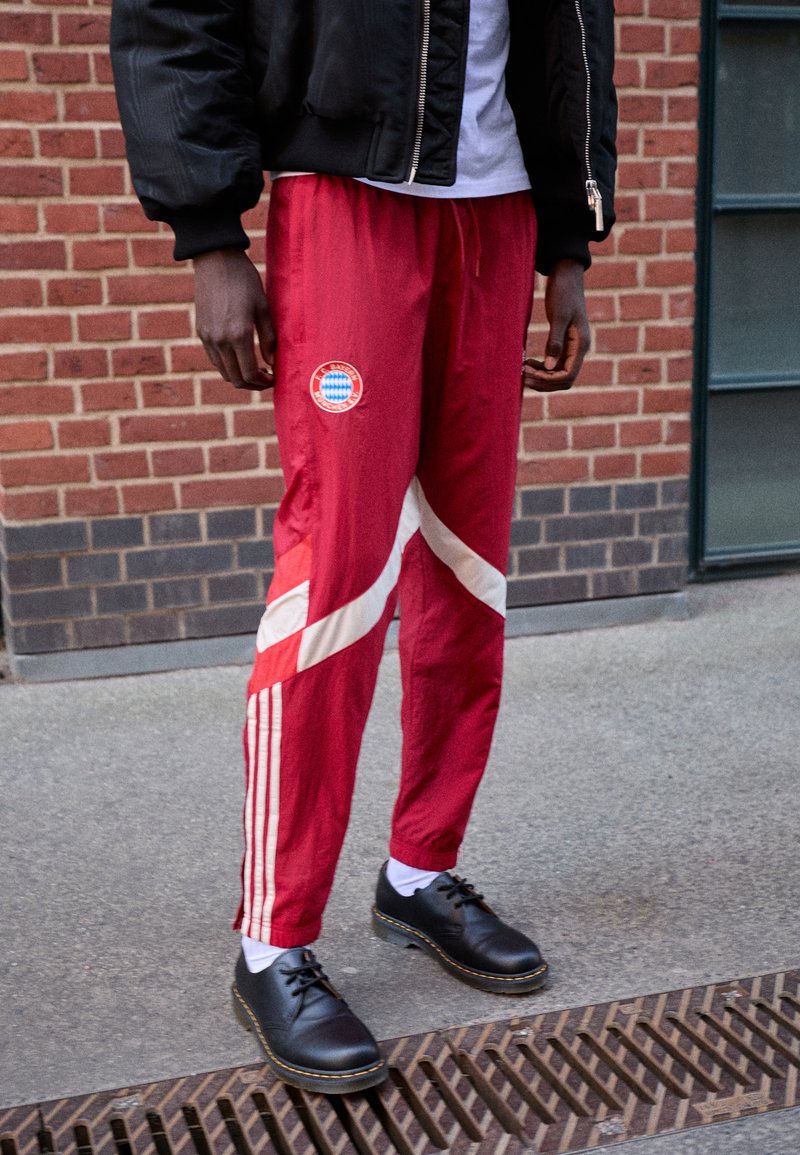 Pantaloni sportivi rossi con strisce diagonali bianche e un logo, realizzati in tessuto leggero, abbinati a scarpe nere e calze bianche.