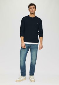 Marinefarbener strukturierter Pullover mit langen Ärmeln, kombiniert mit hellblauen Jeans und beigen Sneakern. Verfügt über ein kleines schwarzes Logo-Detail auf dem Pullover.