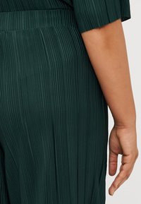 Gros plan du dos d'une femme, portant un vêtement plissé de couleur vert foncé. Une main partiellement visible repose le long de la taille du vêtement.