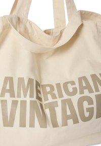 Sac fourre-tout en toile couleur blanc cassé avec un grand texte beige audacieux indiquant "AMERICAN VINTAGE." Doté de doubles poignées et d'une texture douce. Design minimaliste.