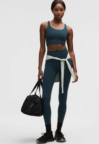 Vihertävä urheiluliivit ja leggingsit, joissa on vaaleanharmaa solmittava vyötäröyksityiskohta. Mukana mustat urheilukengät ja musta duffel-laukku.