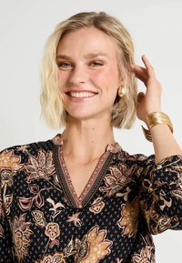 Blouse noire à fleurs avec des accents rose clair et or. Encolure en V avec une bordure décorative. Manches longues avec des poignets à boutons. Texture lisse.
