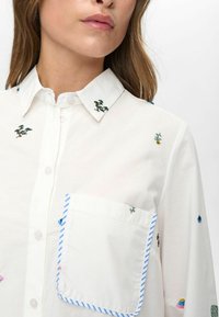 Vrouw in een wit overhemd met knopen, bedekt met kleine geborduurde planten en een borstzakje omlijnd met blauwe en witte strepen.