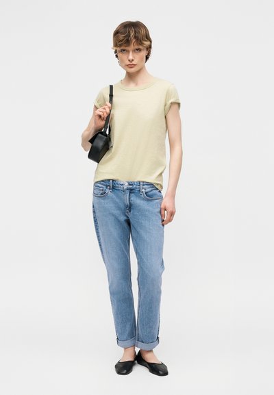 Νεαρό άτομο με κοντά μαλλιά, φοράει μπεζ κοντομάνικο μπλουζάκι με γυρισμένα μανίκια, τζιν ανοιχτού μπλε χρώματος με γυρισμένα τελειώματα, μαύρα μπαλαρίνες και κρατά μια μαύρη τσάντα ώμου.