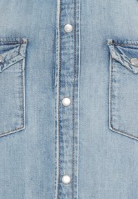 Camicia in denim blu chiaro con due tasche sul petto, bottoni a pressione e cuciture a contrasto lungo i bordi, con una texture leggermente sbiadita.