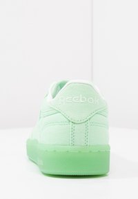 Grön textilskor med en vit vadderad krage, med en genomskinlig grön sula och präglad logotyp på hälen. Slät, strukturerad yta.