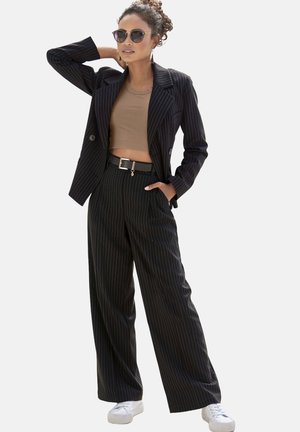 Schwarze Anzugjacke mit Nadelstreifen und Revers, kombiniert mit einem taillierten braunen Crop-Top und weit geschnittenen Hosen. Akzente setzen ein silberner Gürtel und weiße Sneaker.