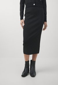 Vrouw in een zwarte midi-jurk met witte stippen, zwarte lange mouwen top en zwarte enkellaarsjes, staand op een effen achtergrond.