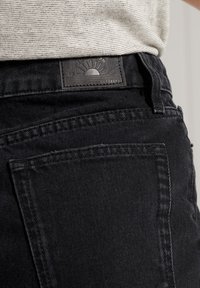 Svarta denimjeans med en lapptag med en sol-logo vid midjan, synliga stygn och en detalj på bakfickan.
