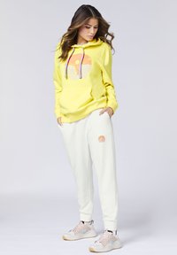 Gelber Hoodie mit einem kreisförmigen Grafikdesign, kombiniert mit cremefarbenen Jogginghosen, die ein kleines Logo tragen. Die Sneaker haben eine hellrosa und graue Farbkombination.