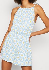 Mono de mezclilla azul con estampado de margaritas en blanco y amarillo, escote halter y tirantes ajustables. Cintura elástica y diseño de pantalones cortos.