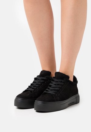 Jambes portant des baskets noires à lacets avec des semelles épaisses en caoutchouc et une doublure intérieure duveteuse sur un fond blanc uni.