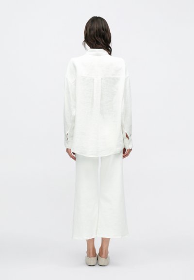 Λευκό λινό πουκάμισο με χαλαρή εφαρμογή, με άνοιγμα στην πλάτη, και λευκό παντελόνι με φαρδιά πόδια. Απλός σχεδιασμός και ουδέτερο χρώμα.