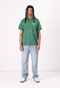 Groene katoenen t-shirt met korte mouwen, ontspannen pasvorm, met een klein logo op de borst. Gecombineerd met lichtblauwe jeans en casual sneakers.