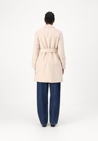 Palton din lână bej cu centură, detaliu pe cusătura din spate și slituri laterale, asortat cu pantaloni din denim întunecat cu croială largă și pantofi negri.