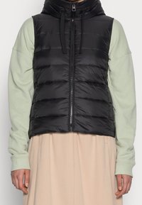 Schwarze Pufferweste mit hohem Kragen, Reißverschluss und horizontalen Nähten. Getragen über einem hellgrünen Langarmshirt. Beige Rock darunter.