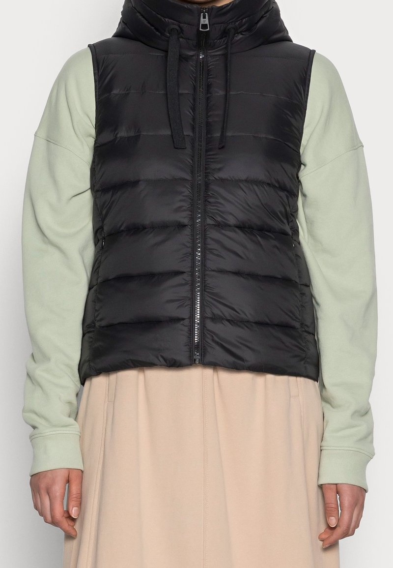 Chaleco acolchado negro con cuello alto, cierre de cremallera y costuras horizontales. Lleva puesto encima de una camisa de manga larga verde claro. Falda beige por debajo.