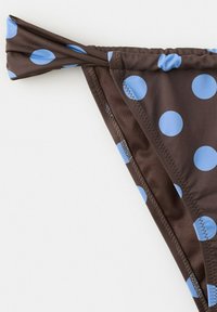 Bas de bikini marron avec de grands pois bleus, doté d'un détail de ficelle nouée sur un côté, sur fond blanc.