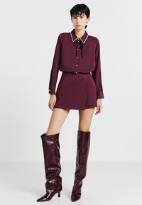 Donna che indossa una blusa viola scuro con dettagli in perle, una gonna avvolgente coordinata e stivali di pelle lucida al ginocchio, in piedi contro uno sfondo bianco.