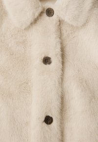 Manteau crémeux en peluche avec une texture douce, dotée d'un col et de quatre boutons foncés sur le devant pour la fermeture.