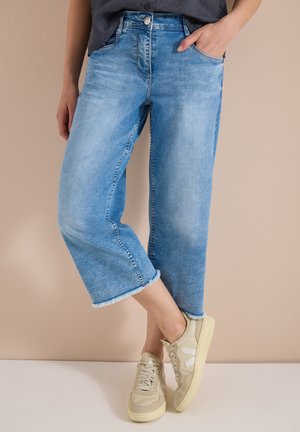 Personne portant un jean large raccourci bleu clair et des baskets beige, debout contre un mur beige, une main dans la poche.