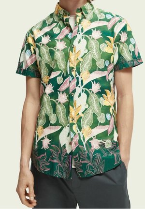 Kortærmet skjorte med knapper med grønne, gule og pink tropiske blade og blomster, båret over en hvid undertrøje.