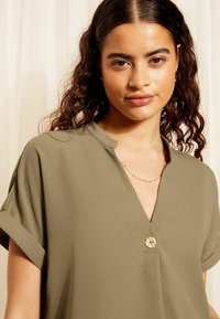 Blusa verde oliva con scollo a V, maniche corte arrotolate e un bottone decorativo dorato frontale, realizzata in un tessuto morbido e leggero.